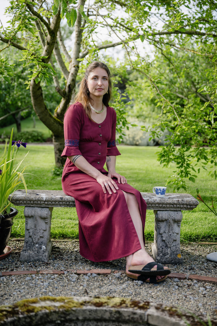 Button front burgundy folklore dress with celtic embroidery styled seated in a garden with front slit and flowing skirt